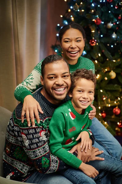 Vertical shot of modern family hugging warmly and smiling at camera with Christmas tree backdrop — Stock Photo