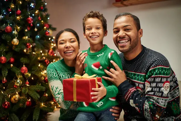 Retrato de la familia afroamericana alegre moderna sosteniendo presente y sonriendo a la cámara, Navidad - foto de stock