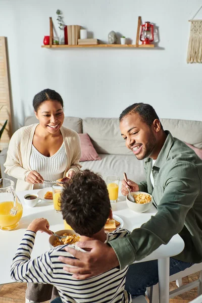 Disparo vertical de la feliz familia afroamericana moderna desayunando y sonriendo alegremente - foto de stock