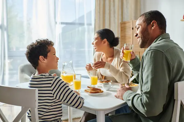 Gioioso adorabile ragazzo afro-americano sorridente a suo padre mentre sua madre distoglie lo sguardo, colazione — Foto stock