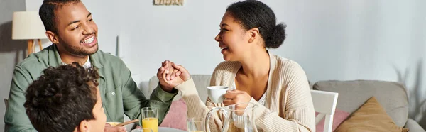 Familia afroamericana feliz en ropa casera casual desayunando y sonriendo el uno al otro, pancarta - foto de stock