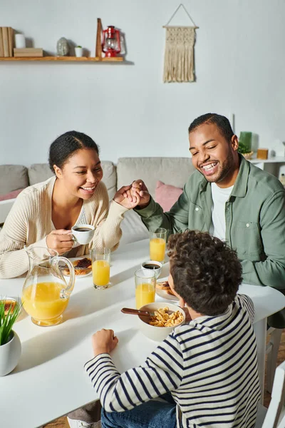 Plano vertical de los padres afroamericanos modernos tomados de la mano en el desayuno y sonriendo a su hijo - foto de stock