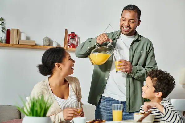 Homme afro-américain joyeux versant du jus d'orange dans un verre tandis que sa femme et son fils lui sourient — Photo de stock