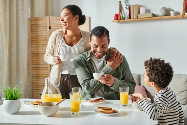 Padres afroamericanos alegres con tazas de café en las manos sonriendo cerca de su hijo desayunando - foto de stock