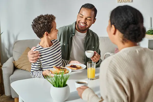 Se centran en el padre y el hijo afroamericanos sonriéndose el uno al otro con la mujer borrosa mirándolos - foto de stock