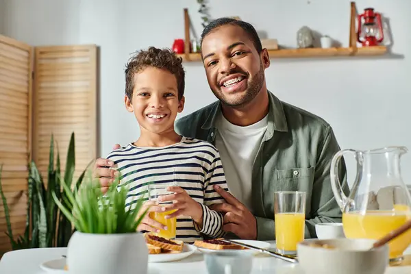 Feliz afroamericano padre e hijo sentado en la mesa del desayuno y sonriendo alegremente a la cámara - foto de stock