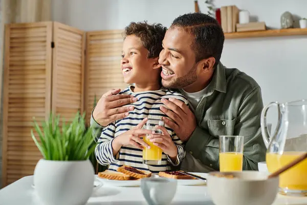 Heureux père afro-américain et fils en tenue de maison décontractée étreignant à la table du petit déjeuner regardant loin — Photo de stock