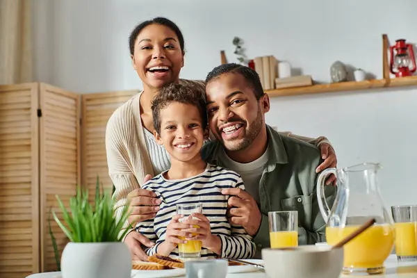 Familia afroamericana alegre abrazándose cálidamente y sonriendo alegremente a la cámara en la mesa del desayuno - foto de stock