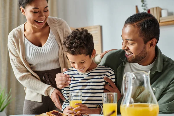 Joyeuse famille afro-américaine moderne étreignant et souriant au petit déjeuner, jus d'orange dans les mains — Photo de stock
