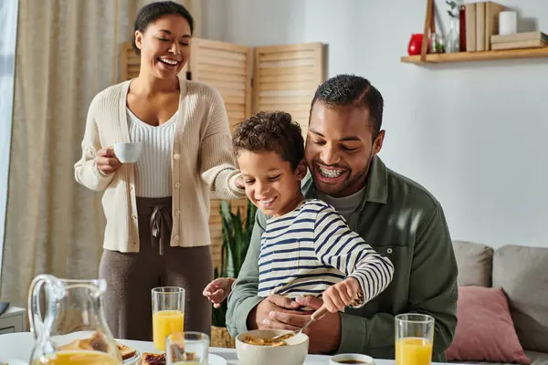 Радісна сучасна афроамериканська сім'я їсть сніданок і п'є каву і апельсиновий сік разом — стокове фото