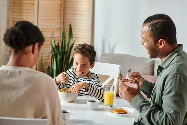 Enfoque en alegre afroamericano chico en ropa de casa disfrutando del desayuno con sus padres borrosos - foto de stock