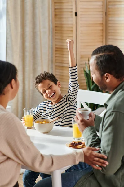 Vertical shot of little african american boy cheering at breakfast while his parent looking at him — Stock Photo