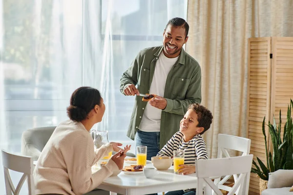 Alegre afroamericano hombre extendiendo mermelada en tostadas y mirando a su esposa e hijo en el desayuno - foto de stock