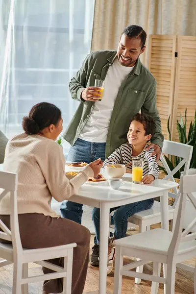 Tiro vertical de feliz afroamericano padre e hijo sonriendo a la madre en la mesa del desayuno - foto de stock