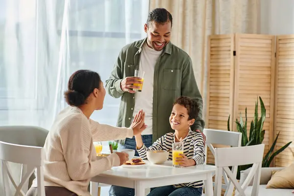 Beautiful modern african american family giving high five at breakfast table, orange juice in hand — Stock Photo