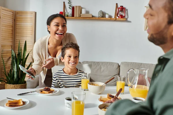 Focus on cheerful african american mother and son in homewear smiling at blurred father at breakfast — Stock Photo