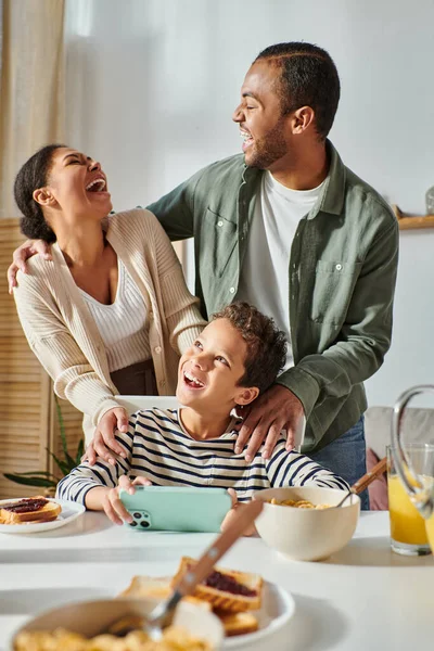 Plan vertical de belle famille afro-américaine riant sincèrement au petit déjeuner, téléphone dans les mains — Photo de stock