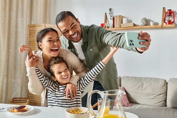 Moderna familia afroamericana alegre tomando selfies en la mesa del desayuno y sonriendo a la cámara - foto de stock