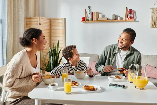 Alegre moderna família afro-americana tomando seu café da manhã e sorrindo um para o outro alegremente — Fotografia de Stock