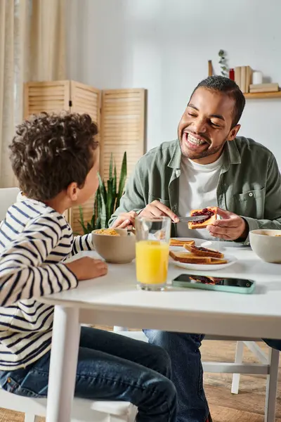 Disparo vertical de niño afroamericano feliz sonriendo a su padre mientras se extiende mermelada en tostadas - foto de stock