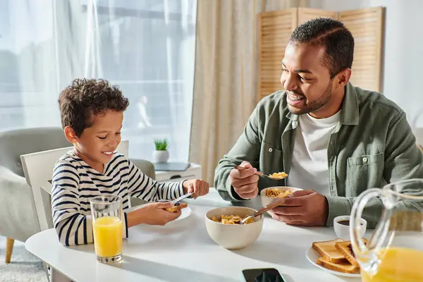 Fröhlich gutaussehender afrikanisch-amerikanischer Vater schaut glücklich zu seinem Sohn, der am Tisch frühstückt — Stockfoto