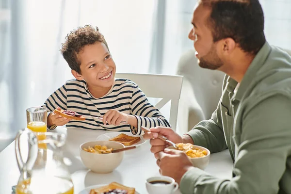 Alegre afro-americano padre e hijo en ropa de casa desayunando y sonriendo el uno al otro - foto de stock