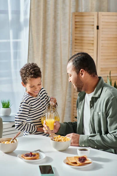 Tiro vertical de pequeno menino afro-americano bonito derramando copo de suco de laranja com sua ajuda pai — Fotografia de Stock