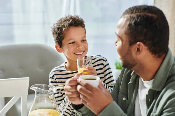 Cheerful adorable african american boy with orange juice smiling at his jolly father at breakfast — Stock Photo