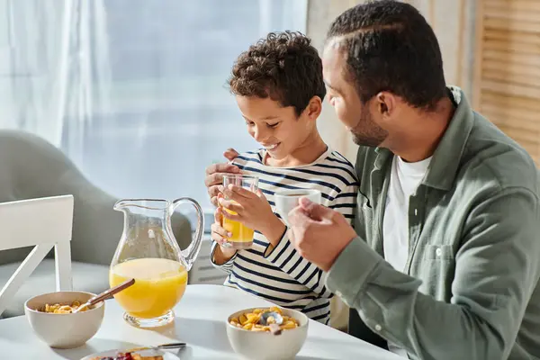 Handsome african american man hugging his son by shoulder and smiling joyfully at breakfast table — Stock Photo