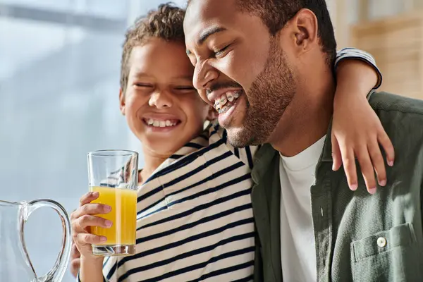 Alegre afroamericano padre e hijo sonriendo felizmente con los ojos cerrados, jugo de naranja en la mano - foto de stock