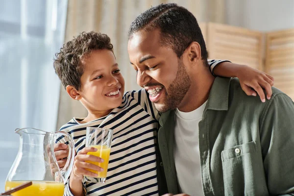 Cheerful african american boy with orange juice hugging his father by shoulder at breakfast table — Stock Photo