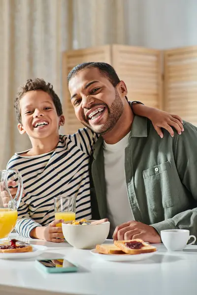 Vertikale Aufnahme eines fröhlichen afrikanisch-amerikanischen Vaters und Sohnes, die sich beim Frühstück umarmen und in die Kamera lächeln — Stockfoto