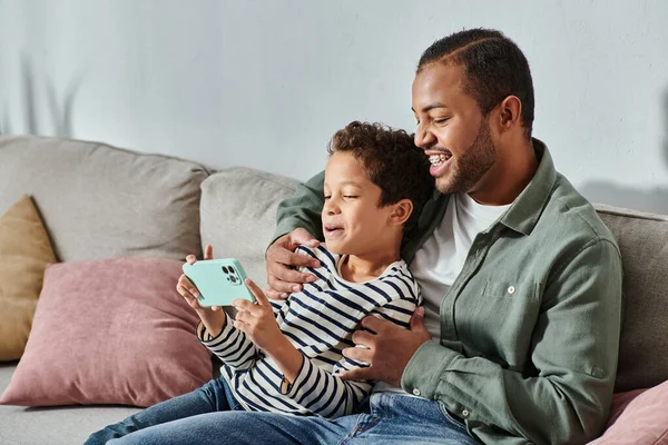 Feliz afroamericano padre e hijo en trajes casuales sentado en el sofá mirando el teléfono móvil - foto de stock