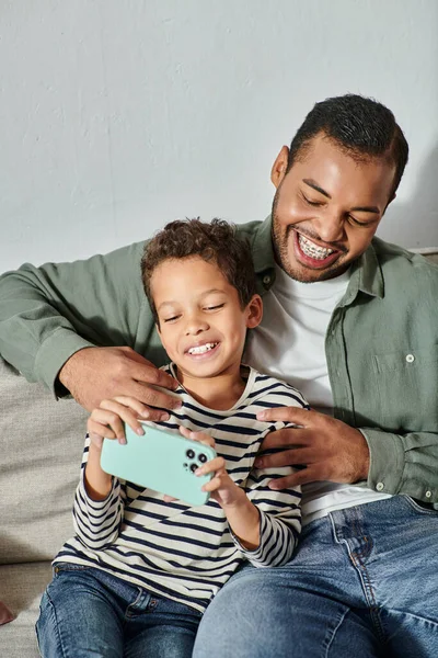 Disparo vertical del alegre padre afroamericano abrazando a su hijo y mirando el teléfono móvil - foto de stock