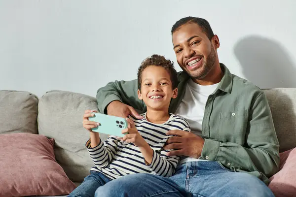 Feliz afroamericano padre e hijo sentado en el sofá con el teléfono en las manos y sonriendo a la cámara - foto de stock