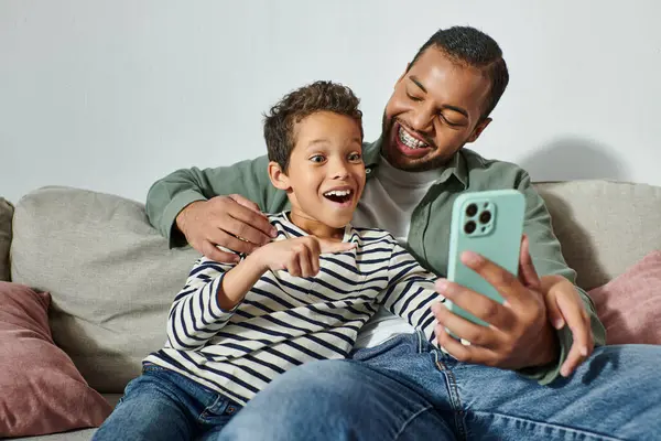 Felice sorpreso ragazzo africano americano guardando sorpreso al telefono cellulare seduto sulle ginocchia del padre — Foto stock