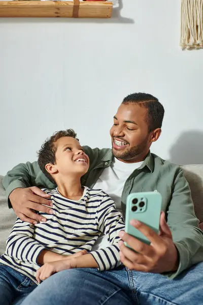 Tiro vertical de menino americano africano alegre sorrindo alegremente para seu pai enquanto toma selfies — Fotografia de Stock