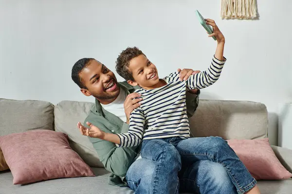 Joyous african american father and son in homewear having fun and taking selfies sitting on sofa — Stock Photo