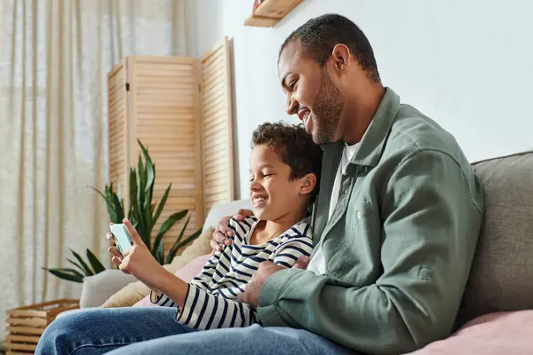 Happy african american father sitting on sofa with his son hugging and smiling at mobile phone — Stock Photo