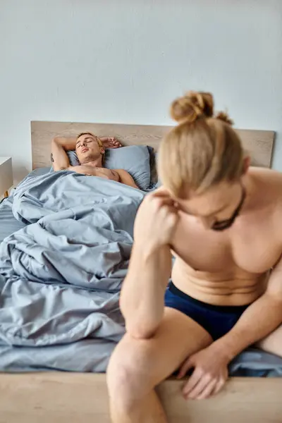 Розчарований бородатий гей чоловік сидить з вклоненою головою біля партнера кохання, спить у спальні, конфлікт — стокове фото