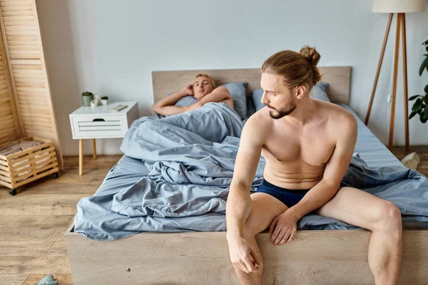 Beleidigter bärtiger Schwuler in Unterhosen, der in der Nähe seines Liebespartners sitzt, der im Schlafzimmer liegt, gestörte Liebe — Stockfoto