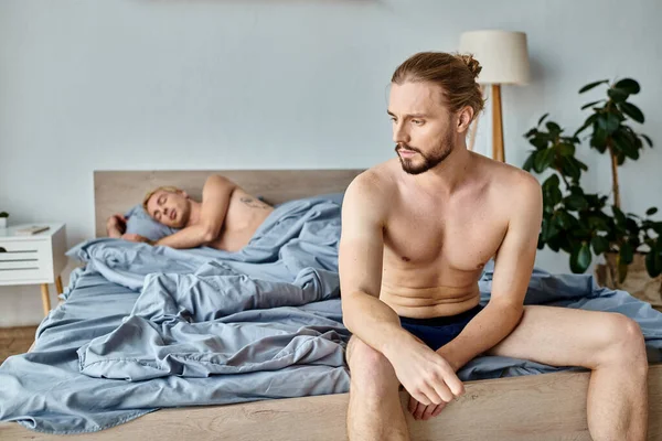 Ofendido barbudo gay hombre en calzoncillos sentado cerca amor socio acostado en dormitorio, problemático amor - foto de stock