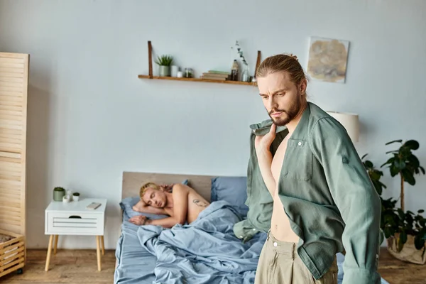 Разочарованный бородатый гей одевается рядом с парнем спит в спальне по утрам, любовный конфликт — стоковое фото