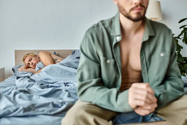 Disappointed gay man lying and looking at boyfriend sitting and dressing up in bedroom, love issues — Stock Photo