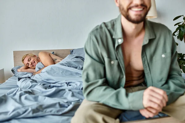 Alegre gay hombre acostado en cama y sonriendo cerca novio sentado en borrosa primer plano, felicidad - foto de stock