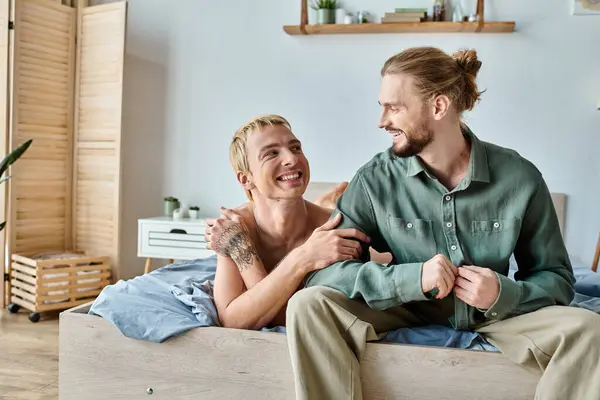 Fröhlich tätowierter schwuler Mann schaut lächelnden bärtigen Freund an, der im Schlafzimmer sitzt, glückliche Beziehung — Stockfoto