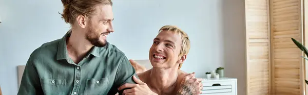 Joyful tattooed gay man looking at smiling bearded boyfriend sitting in bedroom, horizontal banner — Stock Photo