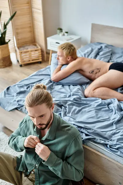 Aufgebracht bärtiger schwuler Mann verkleidet sich in der Nähe beleidigter Freund, der morgens im Schlafzimmer liegt, Liebesprobleme — Stockfoto