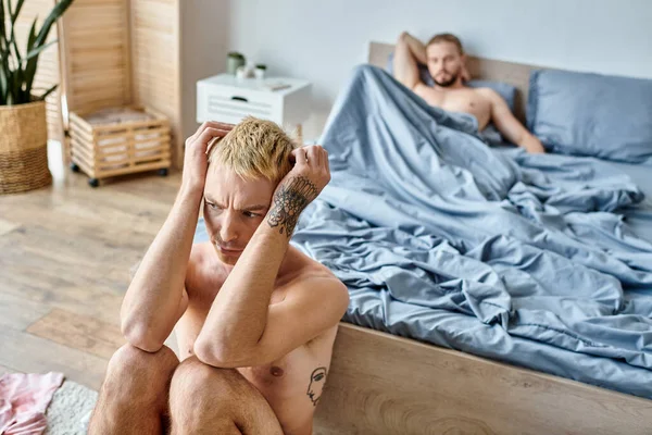 Розчарований татуйований гей чоловік, що сидить поруч з партнером по любові, лежить в спальні вранці, неспокійне кохання — стокове фото