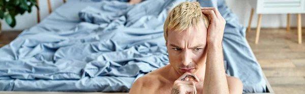 Nachdenklicher und einsamer tätowierter Mann mit blond gefärbten Haaren in der Nähe eines großen Bettes im Schlafzimmer zu Hause, Banner — Stockfoto